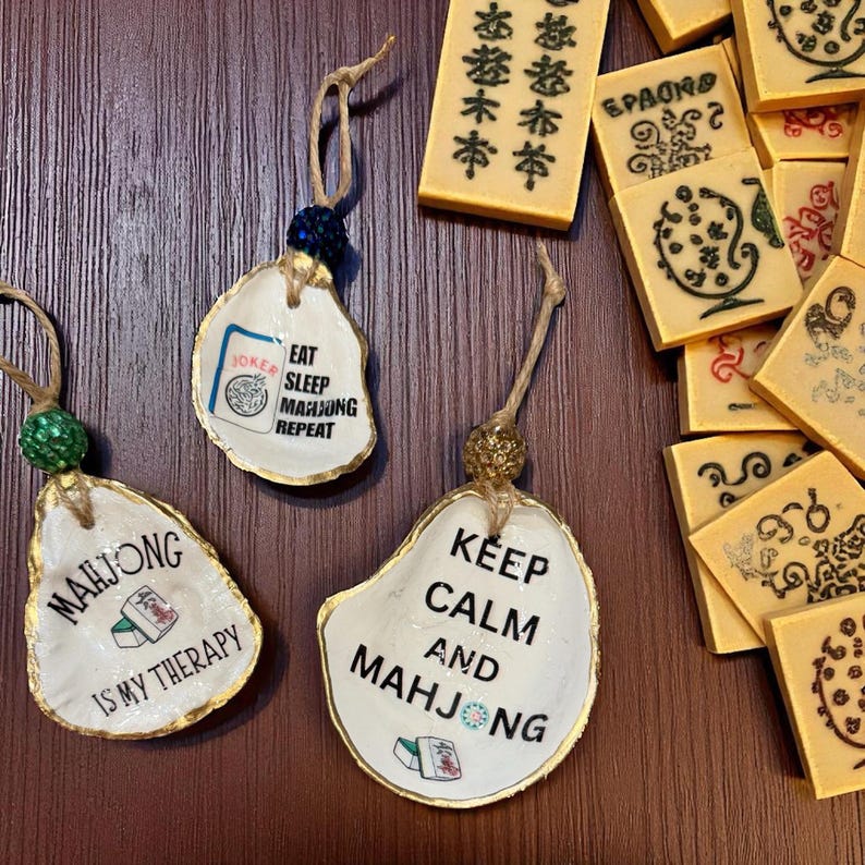 Peut inclure: Ornements faits &agrave; la main en coquille d'hu&icirc;tre avec bordure dor&eacute;e et les phrases "Mahjong is my therapy", "Eat Sleep Mahjong Repeat", et "Keep Calm and Mahjong". Des tuiles de Mahjong sont &eacute;galement visibles.