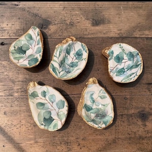 May include: Five decorative oyster shells with gold edges and a watercolor eucalyptus leaf design. The shells are white with green and brown leaf details. The shells are arranged on a wooden surface.