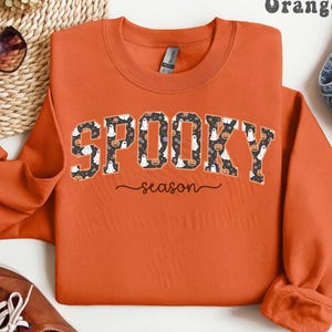 Orange sweatshirt with spooky season design featuring ghosts, pumpkins, and flowers, perfect for fall outfits or Halloween gift.