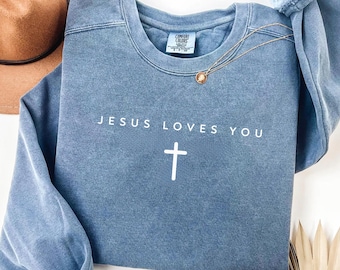 May include: Blue jean-colored sweatshirt with the text "JESUS LOVES YOU" and a white cross. A gold necklace with a pendant is draped on the sweatshirt. A brown hat and dried plant are in the background.