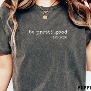 May include: Dark gray t-shirt with the phrase "be pretti good" and the years "1988-2026" printed in white. The shirt has short sleeves and a crew neck. A gold necklace is visible.