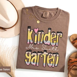 May include: Espresso-colored t-shirt with the words "Kinder Mrs. Smith Garten" in a pencil-themed design. A pink heart is above the word "Kinder". A beige hat and brown sandals are also in the image.