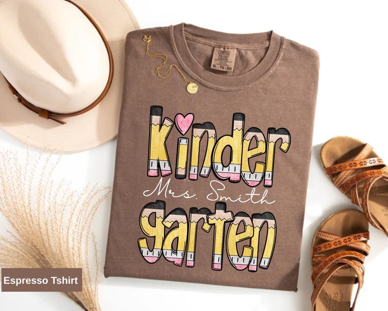 May include: Espresso-colored t-shirt with the words "Kinder Mrs. Smith Garten" in a pencil-themed design. A pink heart is above the word "Kinder". A beige hat and brown sandals are also in the image.