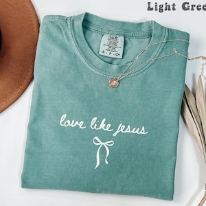 May include: Light green t-shirt with the text "love like jesus" in white script and a bow graphic. A silver necklace with a round pendant rests on the shirt. The shirt is folded and displayed with a brown hat and dried leaves.