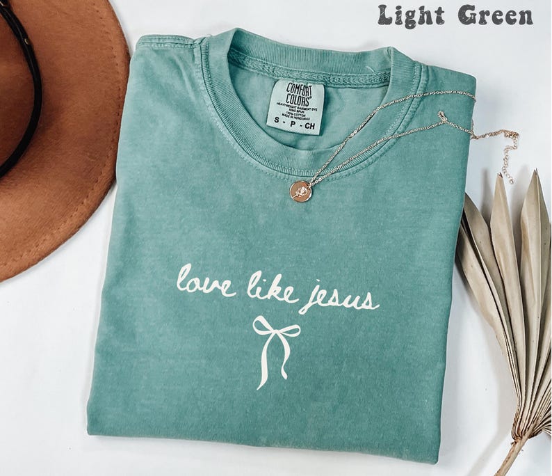 May include: Light green t-shirt with the text "love like jesus" in white script and a bow graphic. A silver necklace with a round pendant rests on the shirt. The shirt is folded and displayed with a brown hat and dried leaves.