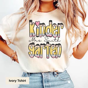 May include: Ivory-colored t-shirt with the words "Kinder Mrs. Smith Garten" in a pencil-themed design. The word "Kinder" has a pink heart above the letter "e". The shirt is made of a soft material.