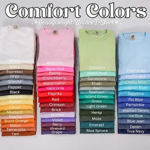 May include: Stacks of folded Comfort Colors heavyweight unisex t-shirts in various colors, including white, pink, green, and blue. Each stack is labeled with the color name, such as "Blossom," "Chambray," and "Black."