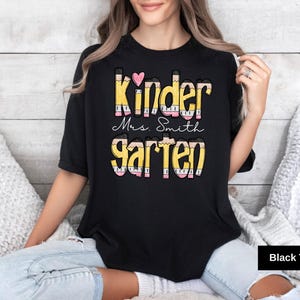 May include: Black t-shirt with the words "Kinder Mrs. Smith Garten" in a colorful pencil-themed design. A pink heart is above the word "Kinder". The shirt is a crew neck style.