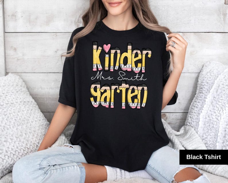May include: Black t-shirt with the words "Kinder Mrs. Smith Garten" in a colorful pencil-themed design. A pink heart is above the word "Kinder". The shirt is a crew neck style.