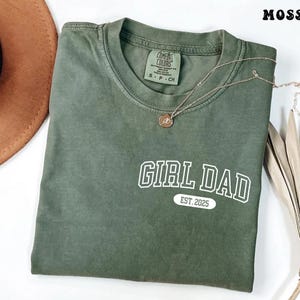 May include: A moss green t-shirt with the text "GIRL DAD EST. 2025" in white. A gold necklace with a pendant is draped across the shirt. A brown hat and dried plant are in the background.