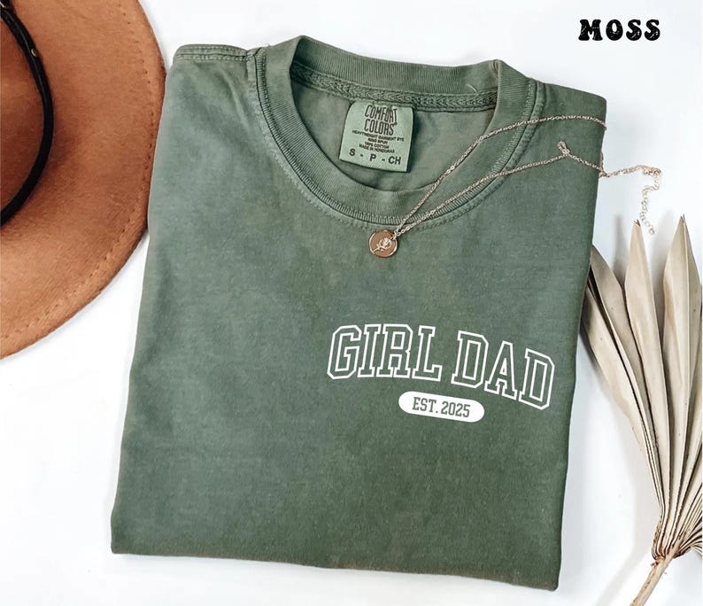 May include: A moss green t-shirt with the text "GIRL DAD EST. 2025" in white. A gold necklace with a pendant is draped across the shirt. A brown hat and dried plant are in the background.