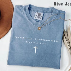 Puede incluir: Una camiseta azul vaquero con el texto "FATHERHOOD IS KINGDOM WORK Proverbs 22:6" y una cruz blanca. Un collar dorado con un colgante descansa sobre la camiseta. La camiseta tiene una etiqueta Comfort Colors.