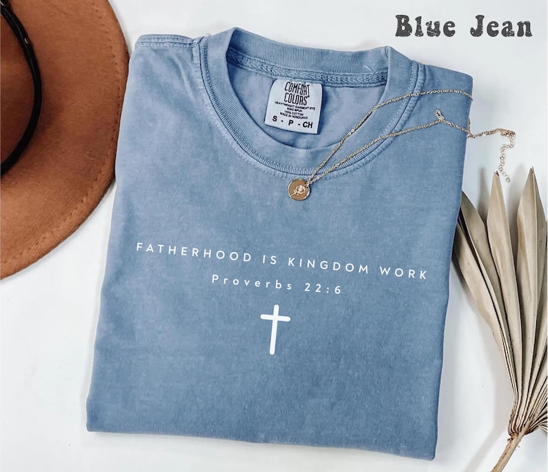 Puede incluir: Una camiseta azul vaquero con el texto "FATHERHOOD IS KINGDOM WORK Proverbs 22:6" y una cruz blanca. Un collar dorado con un colgante descansa sobre la camiseta. La camiseta tiene una etiqueta Comfort Colors.