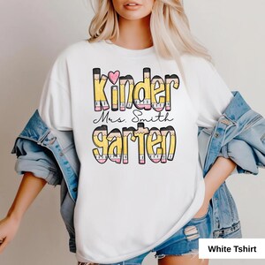 May include: White t-shirt with the words "Kinder Mrs. Smith Garten" in a colorful pencil-themed design. The design includes a pink heart and is suitable for kindergarten teachers. The shirt is made of a soft material.