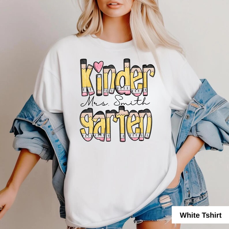 May include: White t-shirt with the words "Kinder Mrs. Smith Garten" in a colorful pencil-themed design. The design includes a pink heart and is suitable for kindergarten teachers. The shirt is made of a soft material.