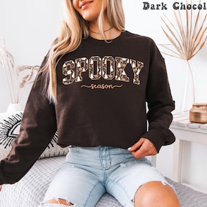 May include: Dark chocolate-colored sweatshirt with the words "SPOOKY season" in a decorative font. The word "SPOOKY" is filled with a pattern of pumpkins and ghosts. The sweatshirt is long-sleeved and has a crew neck.