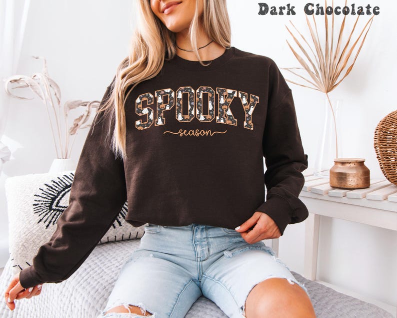 May include: Dark chocolate-colored sweatshirt with the words "SPOOKY season" in a decorative font. The word "SPOOKY" is filled with a pattern of pumpkins and ghosts. The sweatshirt is long-sleeved and has a crew neck.