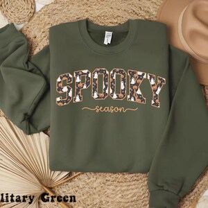 May include: Military green sweatshirt with the words "Spooky season" in a patterned design featuring ghosts and pumpkins. The sweatshirt has long sleeves and a crew neck, perfect for Halloween.