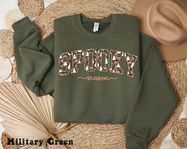 May include: Military green sweatshirt with the words "Spooky season" in a patterned design featuring ghosts and pumpkins. The sweatshirt has long sleeves and a crew neck, perfect for Halloween.