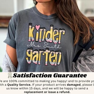 May include: A gray t-shirt with the words "Kinder Mrs. Smith Garten" in a colorful pencil-themed design. The word "Kinder" has a pink heart above it. The shirt is a comfortable, casual style.
