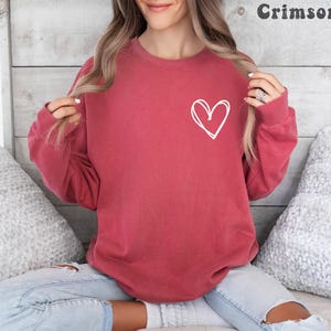 Puede incluir: Sudadera de color carmesí con un contorno de corazón blanco en la parte delantera. La parte superior de manga larga está hecha de un material suave. La palabra "Crimson" está impresa en la esquina superior derecha.