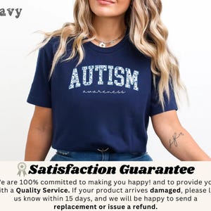 May include: Navy blue t-shirt with the word "AUTISM" in white, floral-patterned letters. The word "awareness" is written below in a script font. The shirt is a crew neck and has short sleeves.