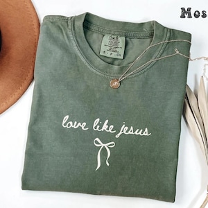 May include: A moss green t-shirt with the phrase "love like jesus" in white script and a bow graphic. A gold necklace with a pendant rests on the shirt. The shirt is folded, and a brown hat is visible in the background.