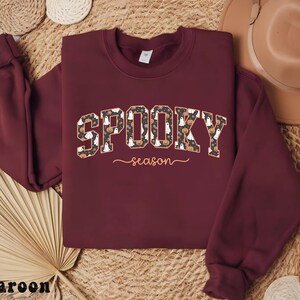 May include: A maroon sweatshirt with the words "SPOOKY season" in a patterned design featuring pumpkins and ghosts. The sweatshirt has long sleeves and a crew neck, perfect for Halloween.