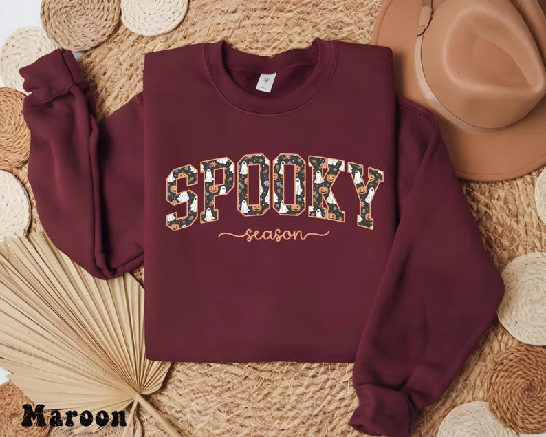 May include: A maroon sweatshirt with the words "SPOOKY season" in a patterned design featuring pumpkins and ghosts. The sweatshirt has long sleeves and a crew neck, perfect for Halloween.