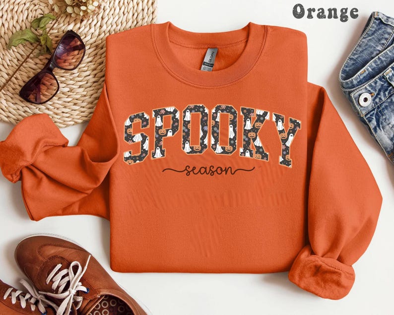 Orange sweatshirt with spooky season design featuring ghosts, pumpkins, and flowers, perfect for fall outfits or Halloween gift.