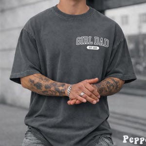May include: A dark gray t-shirt with the text "GIRL DAD EST. 2025" in white. The shirt has a vintage, washed look and a relaxed fit. The sleeves are short, and the shirt is made of a soft material.