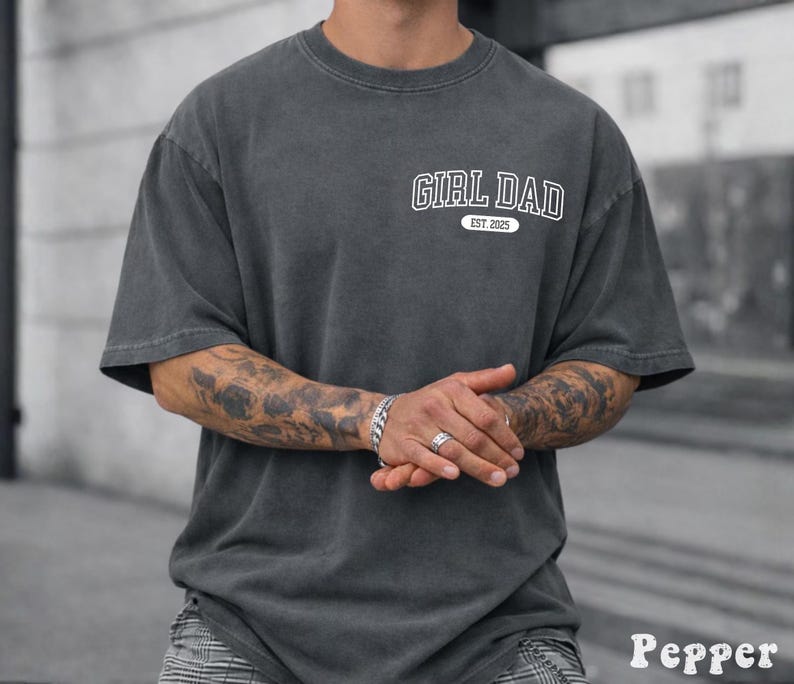 May include: A dark gray t-shirt with the text "GIRL DAD EST. 2025" in white. The shirt has a vintage, washed look and a relaxed fit. The sleeves are short, and the shirt is made of a soft material.