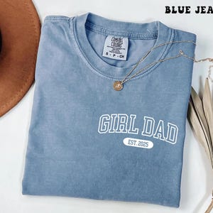 May include: A blue jean Comfort Colors t-shirt with the text "GIRL DAD EST. 2025" in white. A gold necklace with a pendant is draped across the shirt. The shirt is folded and has a tag at the neckline.