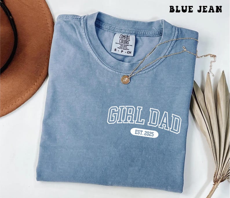 May include: A blue jean Comfort Colors t-shirt with the text "GIRL DAD EST. 2025" in white. A gold necklace with a pendant is draped across the shirt. The shirt is folded and has a tag at the neckline.
