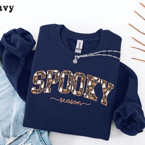 May include: Navy blue sweatshirt with the words "SPOOKY season" in a decorative font. The word "SPOOKY" is filled with a pattern of ghosts and pumpkins. A silver necklace is draped over the sweatshirt.