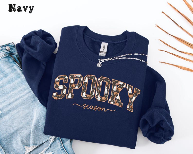May include: Navy blue sweatshirt with the words "SPOOKY season" in a decorative font. The word "SPOOKY" is filled with a pattern of ghosts and pumpkins. A silver necklace is draped over the sweatshirt.