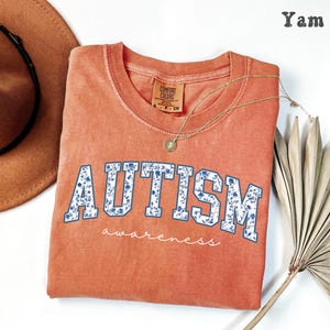 May include: A coral-colored Comfort Colors crew neck t-shirt with the word "AUTISM" in white block letters filled with a blue floral pattern. The word "awareness" is written below in a script font. A gold necklace and a brown hat are also in the image.