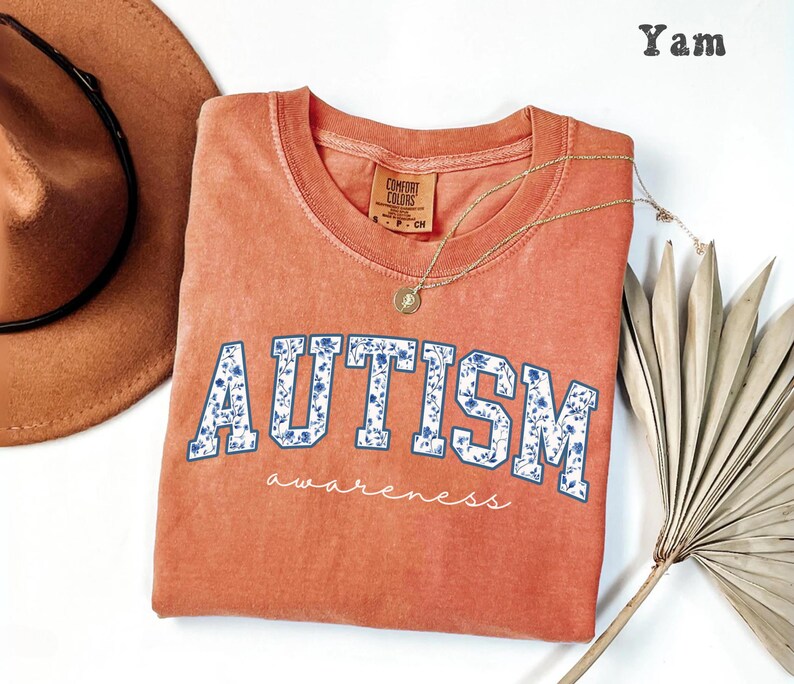 May include: A coral-colored Comfort Colors crew neck t-shirt with the word "AUTISM" in white block letters filled with a blue floral pattern. The word "awareness" is written below in a script font. A gold necklace and a brown hat are also in the image.