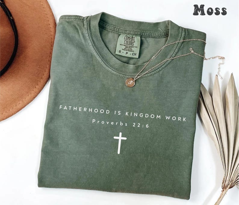 Puede incluir: Una camiseta verde oliva con el texto "FATHERHOOD IS KINGDOM WORK Proverbs 22:6" y una cruz blanca. Un collar dorado con un colgante descansa sobre la camiseta. Un sombrero marr&oacute;n y hojas de palma secas est&aacute;n en el fondo.