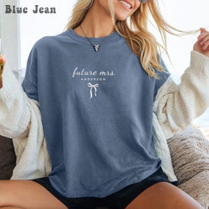 Puede incluir: Una camiseta azul vaquero con el texto "future mrs. Anderson" y un lazo blanco. La camiseta tiene cuello redondo y mangas cortas. La camiseta es de corte holgado.