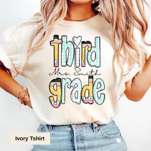 May include: Ivory-colored t-shirt with the words "Third Grade Mrs. Smith" in a colorful, playful font. The design includes pencil and ruler accents, perfect for teachers or students. The shirt is made of a soft material.