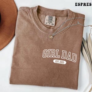 May include: Espresso-colored t-shirt with the text "GIRL DAD EST. 2025" in white. The shirt has a crew neck and a small tag on the collar. A silver necklace with a pendant is draped across the shirt.