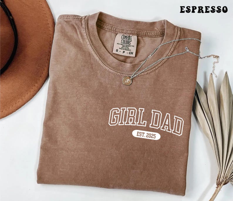 May include: Espresso-colored t-shirt with the text "GIRL DAD EST. 2025" in white. The shirt has a crew neck and a small tag on the collar. A silver necklace with a pendant is draped across the shirt.