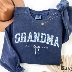 May include: Navy blue sweatshirt with the word "GRANDMA" in white block letters filled with a blue floral pattern. Below the word "GRANDMA" is the text "EST. 2026" and a white bow graphic.