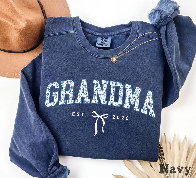 May include: Navy blue sweatshirt with the word "GRANDMA" in white block letters filled with a blue floral pattern. Below the word "GRANDMA" is the text "EST. 2026" and a white bow graphic.