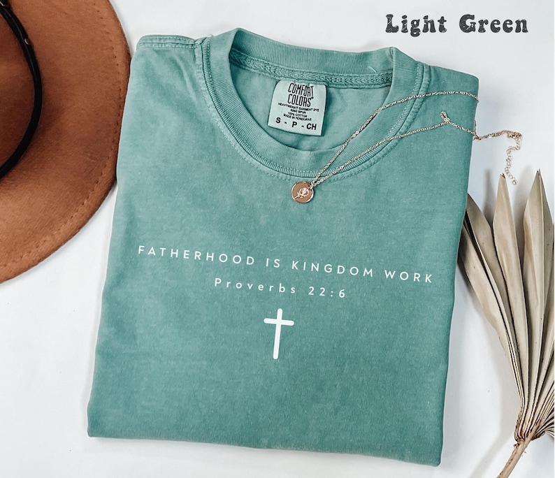 Puede incluir: Camiseta verde claro con el texto "FATHERHOOD IS KINGDOM WORK Proverbs 22:6" y una cruz blanca. Un collar plateado con un colgante circular descansa sobre la camiseta. Un sombrero marr&oacute;n es visible en el fondo.
