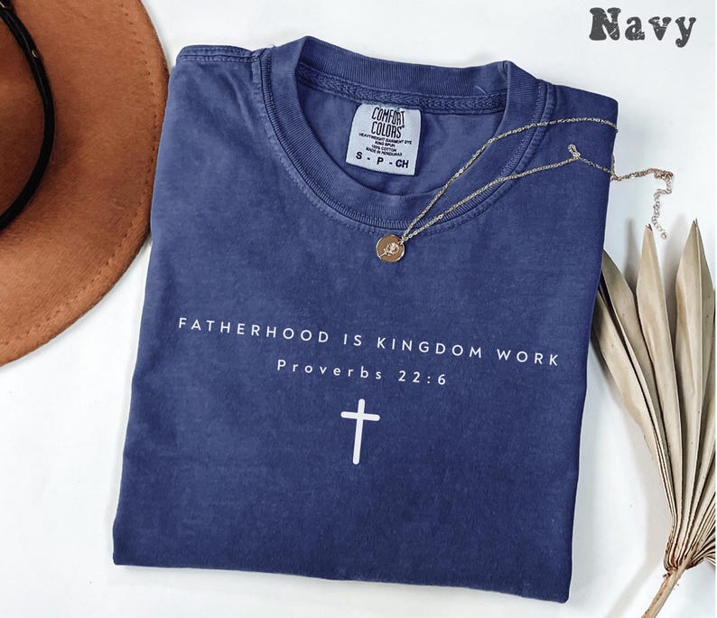 Puede incluir: Camiseta azul marino con el texto "FATHERHOOD IS KINGDOM WORK Proverbs 22:6" y una cruz blanca. Un collar dorado con un colgante descansa sobre la camiseta. La camiseta est&aacute; doblada sobre una superficie blanca.