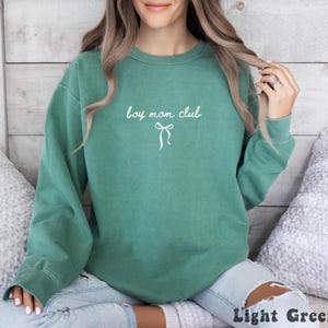 K&ouml;nnte beinhalten: Hellgr&uuml;nes Sweatshirt mit dem wei&szlig;en Schriftzug "boy mom club" und einer wei&szlig;en Schleifen-Grafik. Das Sweatshirt hat einen Rundhalsausschnitt und lange &Auml;rmel. Unten rechts im Bild steht der Text "Light Green".