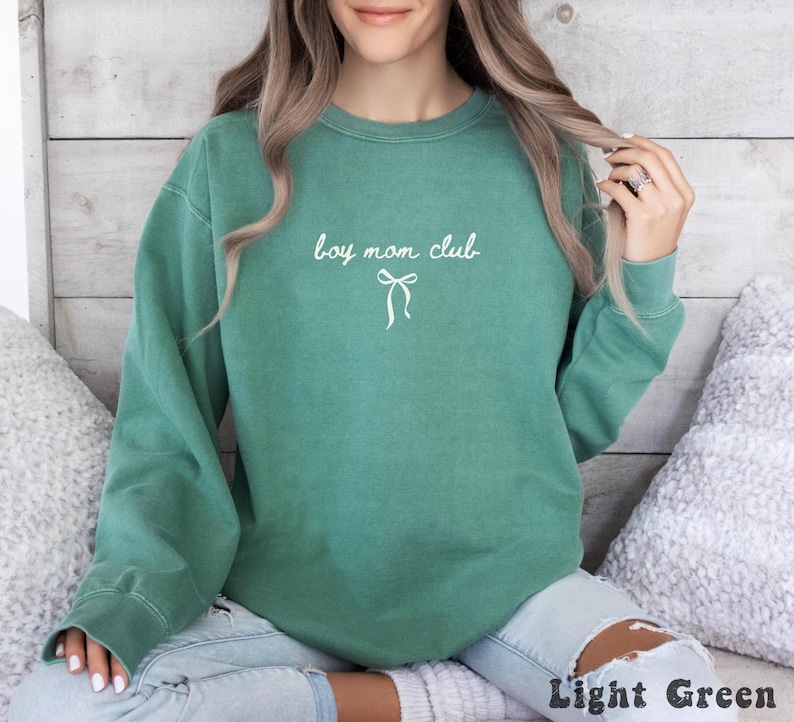 K&ouml;nnte beinhalten: Hellgr&uuml;nes Sweatshirt mit dem wei&szlig;en Schriftzug "boy mom club" und einer wei&szlig;en Schleifen-Grafik. Das Sweatshirt hat einen Rundhalsausschnitt und lange &Auml;rmel. Unten rechts im Bild steht der Text "Light Green".