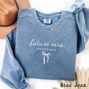 Puede incluir: Una sudadera de cuello redondo color azul jean con el texto en blanco future mrs. ANDERSON sobre un gráfico de lazo blanco. La prenda tiene una etiqueta Comfort Colors en el cuello interior y se acompaña de un collar de cadena dorada.